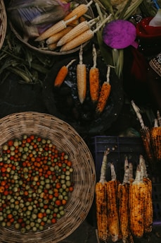 An assortment of fresh produce is arranged in a market setting. There are multiple ears of corn, both raw and grilled, displayed in baskets and on a rustic surface. One basket contains small, round fruit or vegetables in shades of green, orange, and red. The setting appears lively and rustic, indicating a market atmosphere.