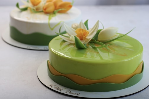Two beautifully crafted cakes with smooth, glossy surfaces are displayed, each adorned with intricate floral decorations. The front cake is primarily green with a wave of yellow accent and features elaborate sugar flowers in white and yellow hues. The background cake has a lighter color scheme with similar floral design elements.
