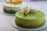 Two beautifully crafted cakes with smooth, glossy surfaces are displayed, each adorned with intricate floral decorations. The front cake is primarily green with a wave of yellow accent and features elaborate sugar flowers in white and yellow hues. The background cake has a lighter color scheme with similar floral design elements.