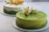 Two beautifully crafted cakes with smooth, glossy surfaces are displayed, each adorned with intricate floral decorations. The front cake is primarily green with a wave of yellow accent and features elaborate sugar flowers in white and yellow hues. The background cake has a lighter color scheme with similar floral design elements.