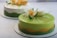 Two beautifully crafted cakes with smooth, glossy surfaces are displayed, each adorned with intricate floral decorations. The front cake is primarily green with a wave of yellow accent and features elaborate sugar flowers in white and yellow hues. The background cake has a lighter color scheme with similar floral design elements.