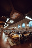 A large library interior filled with rows of wooden tables and chairs, occupied by numerous people engaged in reading and studying. The ceiling is high with ornate detailing and features several large circular chandeliers. Bookshelves line the walls, and the space is illuminated warmly by overhead lights and lamps on each study table.
