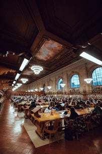 A large library interior filled with rows of wooden tables and chairs, occupied by numerous people engaged in reading and studying. The ceiling is high with ornate detailing and features several large circular chandeliers. Bookshelves line the walls, and the space is illuminated warmly by overhead lights and lamps on each study table.