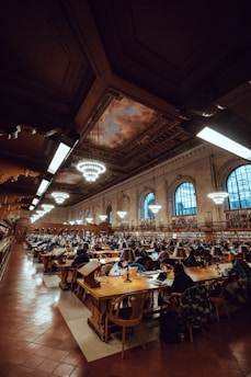 A large library interior filled with rows of wooden tables and chairs, occupied by numerous people engaged in reading and studying. The ceiling is high with ornate detailing and features several large circular chandeliers. Bookshelves line the walls, and the space is illuminated warmly by overhead lights and lamps on each study table.