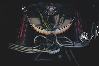 A close-up view of a car engine with a chrome air filter cover in the center. The engine features red accents and visible wiring and hoses surrounding the central component.
