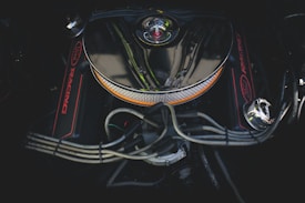 A close-up view of a car engine with a chrome air filter cover in the center. The engine features red accents and visible wiring and hoses surrounding the central component.