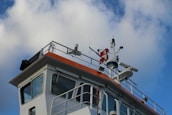 A close-up view of a ship's bridge featuring various navigational equipment, antennas, and instruments. The structure is painted white with an orange trim. The sky in the background is blue with scattered clouds.