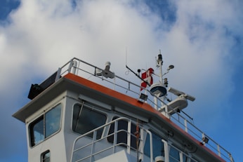 A close-up view of a ship's bridge featuring various navigational equipment, antennas, and instruments. The structure is painted white with an orange trim. The sky in the background is blue with scattered clouds.
