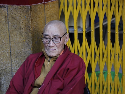 An elderly person with glasses and a shaved head, wearing a red robe, is seated against a backdrop of a yellow metal gate. The setting includes a textured stone wall and the person appears contemplative.