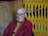 An elderly person with glasses and a shaved head, wearing a red robe, is seated against a backdrop of a yellow metal gate. The setting includes a textured stone wall and the person appears contemplative.