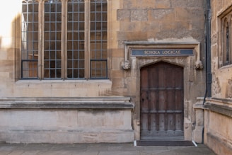 An old stone building facade featuring a large wooden door with a sign above it reading 'Schola Logicae'. The door is framed by two carved stone heads. Next to the door are tall, multi-paned windows with a Gothic architectural style.