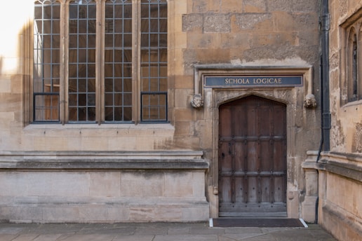 An old stone building facade featuring a large wooden door with a sign above it reading 'Schola Logicae'. The door is framed by two carved stone heads. Next to the door are tall, multi-paned windows with a Gothic architectural style.