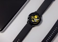 A black smartwatch with a round face displaying a digital interface. The screen shows a heart rate, date, and steps count in bright yellow text against a dark background. Nearby, a black watch strap with multiple holes is visible along with a corner of a sleek black electronic device.