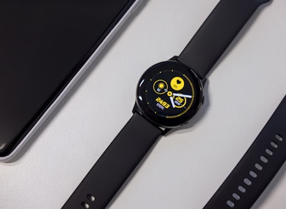 A black smartwatch with a round face displaying a digital interface. The screen shows a heart rate, date, and steps count in bright yellow text against a dark background. Nearby, a black watch strap with multiple holes is visible along with a corner of a sleek black electronic device.