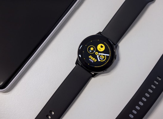 A black smartwatch with a round face displaying a digital interface. The screen shows a heart rate, date, and steps count in bright yellow text against a dark background. Nearby, a black watch strap with multiple holes is visible along with a corner of a sleek black electronic device.