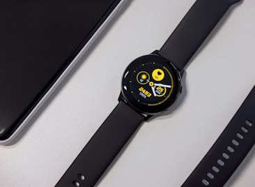 A black smartwatch with a round face displaying a digital interface. The screen shows a heart rate, date, and steps count in bright yellow text against a dark background. Nearby, a black watch strap with multiple holes is visible along with a corner of a sleek black electronic device.