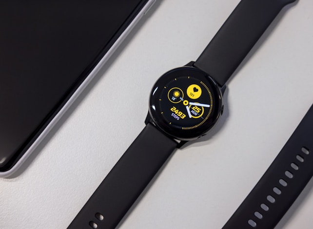 A black smartwatch with a round face displaying a digital interface. The screen shows a heart rate, date, and steps count in bright yellow text against a dark background. Nearby, a black watch strap with multiple holes is visible along with a corner of a sleek black electronic device.