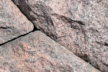 A close-up view of rough, textured granite stones with visible grain and natural color variations. The stones are arranged with visible gaps in between, highlighting their irregular, natural formation.