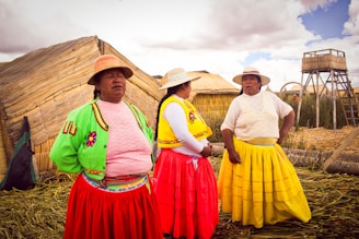 Three women dressed in vibrant traditional clothing stand in front of reed houses. They are wearing colorful skirts and sweaters with intricate designs. The background shows a rural setting with thatched structures and a tall reed hut.