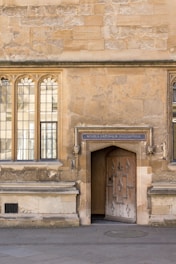 An old stone building with arched windows and an ornate wooden door. Above the door is a sign that reads 'Schola Naturalis Philosophiae'. The wall has a textured, weathered appearance, and the ground is paved with stones.