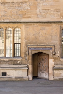 An old stone building with arched windows and an ornate wooden door. Above the door is a sign that reads 'Schola Naturalis Philosophiae'. The wall has a textured, weathered appearance, and the ground is paved with stones.