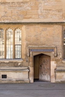 An old stone building with arched windows and an ornate wooden door. Above the door is a sign that reads 'Schola Naturalis Philosophiae'. The wall has a textured, weathered appearance, and the ground is paved with stones.