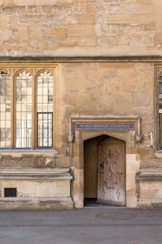 An old stone building with arched windows and an ornate wooden door. Above the door is a sign that reads 'Schola Naturalis Philosophiae'. The wall has a textured, weathered appearance, and the ground is paved with stones.