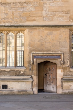 An old stone building with arched windows and an ornate wooden door. Above the door is a sign that reads 'Schola Naturalis Philosophiae'. The wall has a textured, weathered appearance, and the ground is paved with stones.