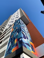 A high-rise building towers upwards with a colorful geometric mural adorning its facade. The mural features abstract shapes in vibrant blues, oranges, and yellows. The sky is clear and blue, providing a striking backdrop to the building's modern design.