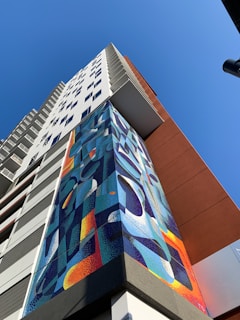 A high-rise building towers upwards with a colorful geometric mural adorning its facade. The mural features abstract shapes in vibrant blues, oranges, and yellows. The sky is clear and blue, providing a striking backdrop to the building's modern design.