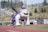 A large inflatable bulldog mascot wearing a crown and cape is set up on a running track. There is a person jogging nearby and some equipment visible. The background features a grassy hill with scattered trees and some buildings in the distance.
