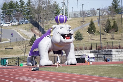 A large inflatable bulldog mascot wearing a crown and cape is set up on a running track. There is a person jogging nearby and some equipment visible. The background features a grassy hill with scattered trees and some buildings in the distance.