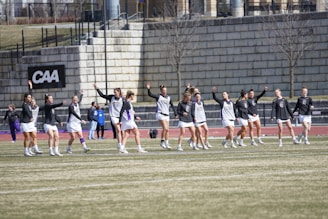 A group of athletes warming up or practicing on a grassy field near a track. They are wearing athletic gear, including shirts and shorts, and appear to be involved in a group exercise or routine. Most have one arm raised, and they are positioned in a line with a large stone wall in the background. A sign on the wall reads 'CAA', indicating a sports conference or event.