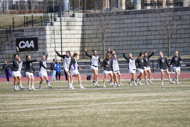 A group of athletes warming up or practicing on a grassy field near a track. They are wearing athletic gear, including shirts and shorts, and appear to be involved in a group exercise or routine. Most have one arm raised, and they are positioned in a line with a large stone wall in the background. A sign on the wall reads 'CAA', indicating a sports conference or event.