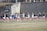A group of athletes warming up or practicing on a grassy field near a track. They are wearing athletic gear, including shirts and shorts, and appear to be involved in a group exercise or routine. Most have one arm raised, and they are positioned in a line with a large stone wall in the background. A sign on the wall reads 'CAA', indicating a sports conference or event.
