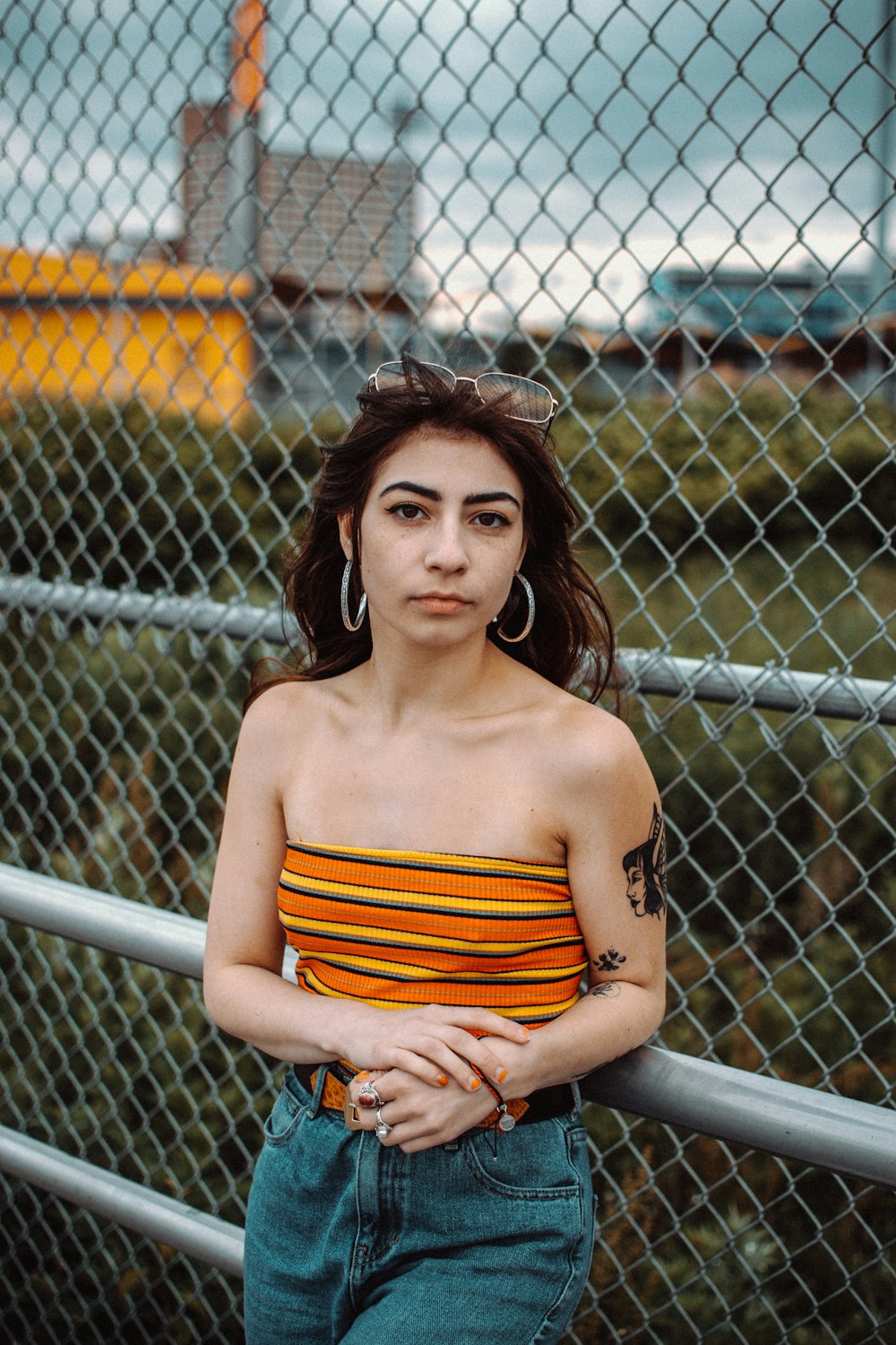 woman wearing orange and yellow stripe off-shoulder and blue jeans lying on cyclone fence