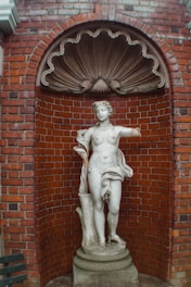 A classical marble statue of a nude female figure with one arm missing stands against a red brick wall. The statue is placed in an arched niche with a decorative shell-like element above. The statue is mounted on a circular pedestal, and there is part of a wooden bench visible on the left side of the image.