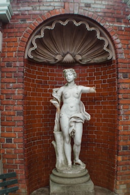 A classical marble statue of a nude female figure with one arm missing stands against a red brick wall. The statue is placed in an arched niche with a decorative shell-like element above. The statue is mounted on a circular pedestal, and there is part of a wooden bench visible on the left side of the image.