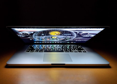 A laptop with a partially closed lid sits on a wooden surface with its screen displaying a car dashboard graphic. The keyboard is visible, illuminated by the light from the screen, creating a dramatic effect against the dark background.