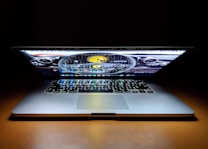 A laptop with a partially closed lid sits on a wooden surface with its screen displaying a car dashboard graphic. The keyboard is visible, illuminated by the light from the screen, creating a dramatic effect against the dark background.