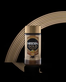 A jar of Nescafé Gold Espresso with a golden lid is displayed against a black background. The jar is centrally positioned, and there are curved golden light reflections behind it that create an elegant arc.