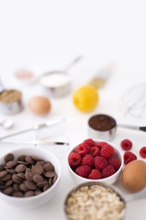 An arrangement of various baking ingredients on a white surface, including a bowl of chocolate chips, a bowl of fresh raspberries, eggs, a lemon, oats, and measuring cups containing flour and cocoa powder. A metal whisk and other baking utensils are also visible.