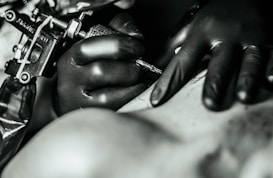 A close-up shot of a tattooing process, featuring a tattoo artist's gloved hands holding a tattoo machine while working on a person's skin.