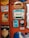A variety of vintage advertisement signs and labels are affixed to a red wall. These include ads for products such as soap, milk, kerosene, and a radio. The designs are colorful and nostalgic, featuring retro typography and illustrations.