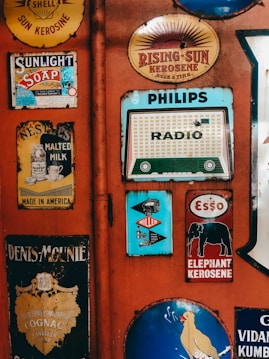A variety of vintage advertisement signs and labels are affixed to a red wall. These include ads for products such as soap, milk, kerosene, and a radio. The designs are colorful and nostalgic, featuring retro typography and illustrations.