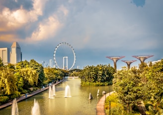 The image features a vibrant cityscape with lush greenery and a large Ferris wheel in the background. Notable are the futuristic tree-like structures that extend above the treetops. A serene waterway with fountains runs through the park, creating a blend of natural and urban elements.