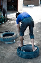A person is bending over a tire on the ground, appearing to be engaged in a manual task. The setting is outdoors, with two tires visible and the ground made up of dirt.