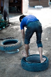 A person is bending over a tire on the ground, appearing to be engaged in a manual task. The setting is outdoors, with two tires visible and the ground made up of dirt.