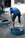 A person is bending over a tire on the ground, appearing to be engaged in a manual task. The setting is outdoors, with two tires visible and the ground made up of dirt.