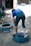 A person is bending over a tire on the ground, appearing to be engaged in a manual task. The setting is outdoors, with two tires visible and the ground made up of dirt.